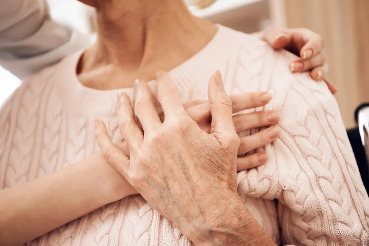 A younger caregiver's hand rests gently on the shoulder of an elderly woman in a soft pink sweater.